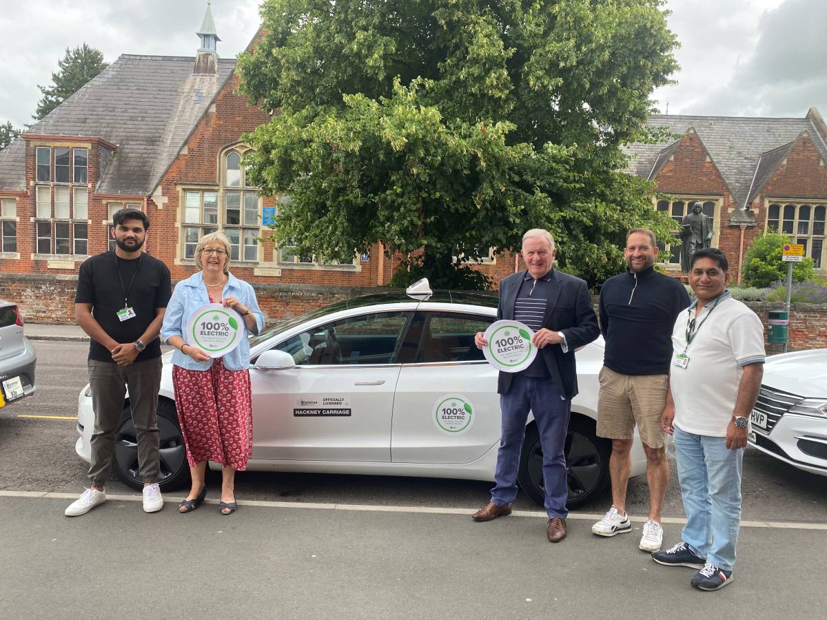 Two district councillors holding electric vehicle branding standing by an electric cab with three drivers.