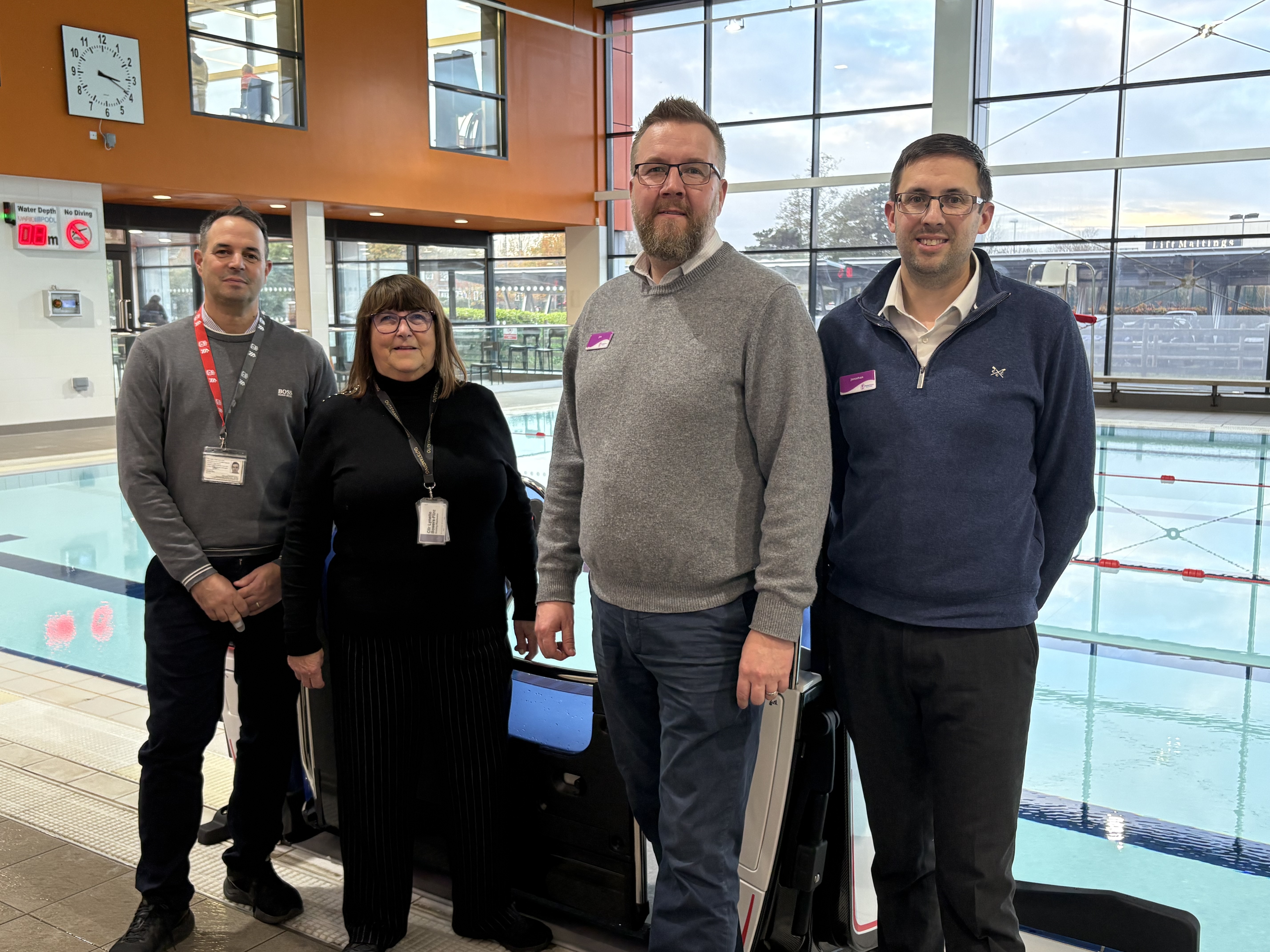 The image features four people looking towards the camera standing in front of a pool pod at a swimming pool