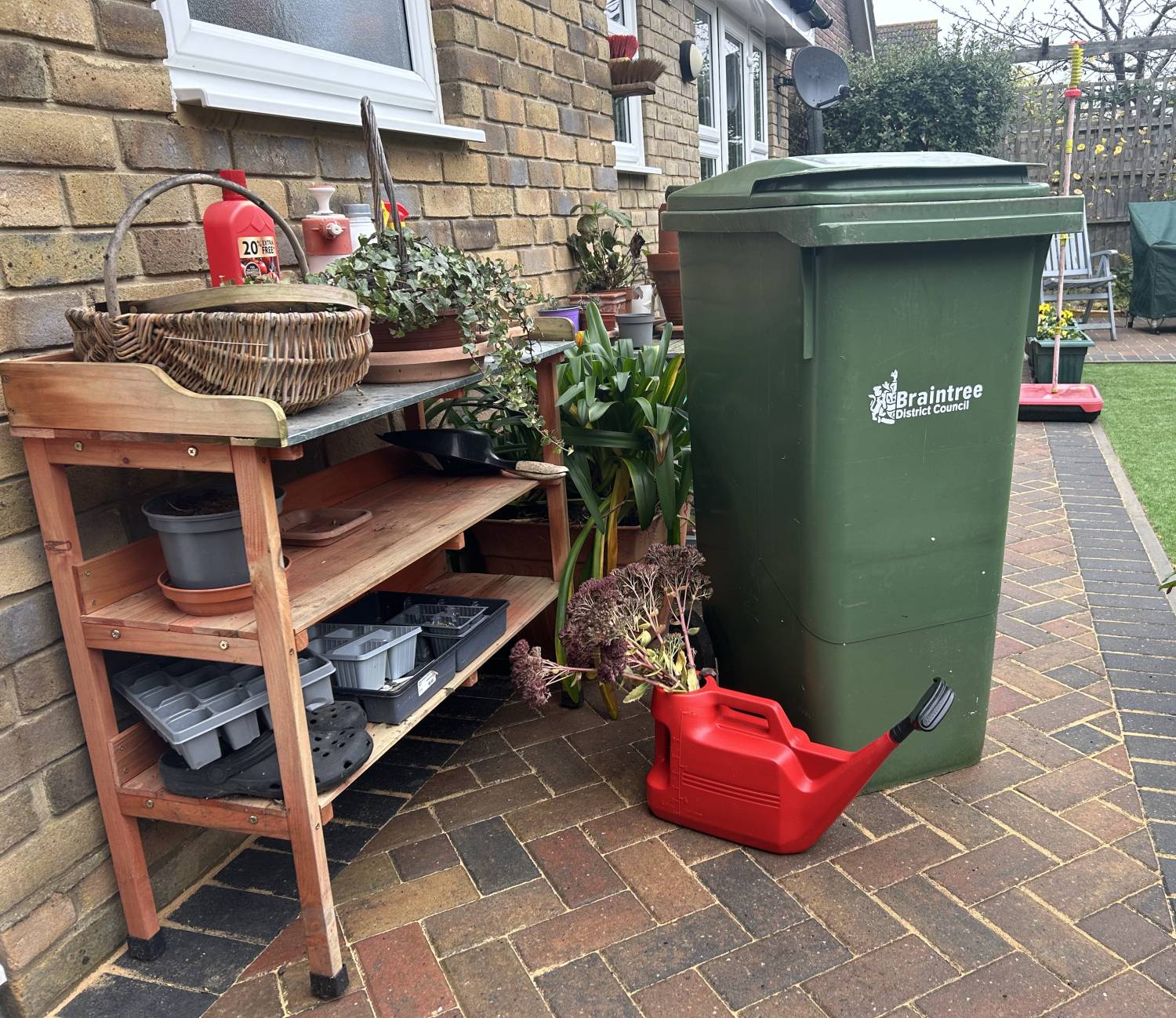 A garden waste bin in a garden, next to shelving with various gardening objects on it