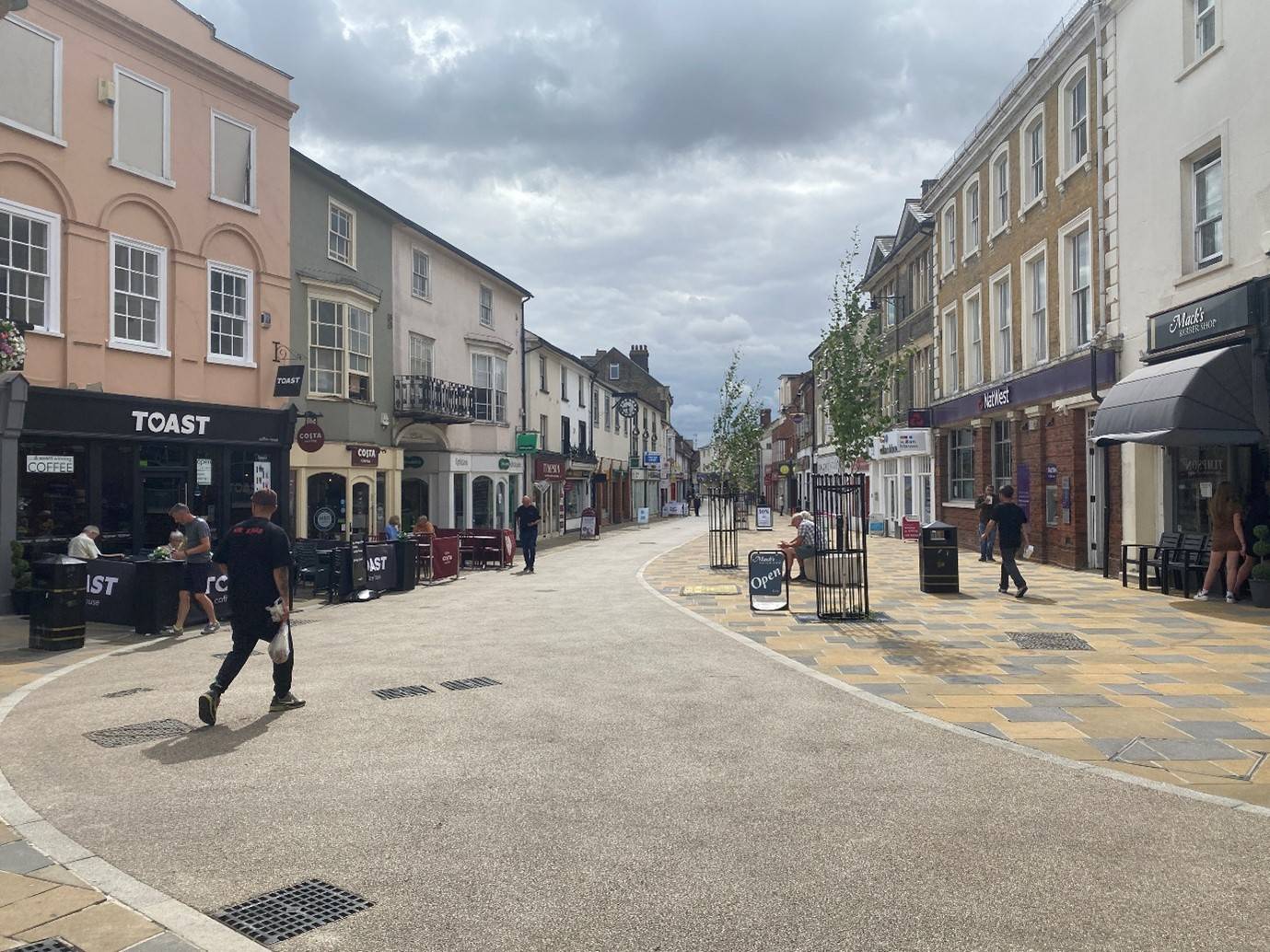 A wide shot of the town centre with buildings on the left and right hand side, people are also sat on chairs and walking down the street.