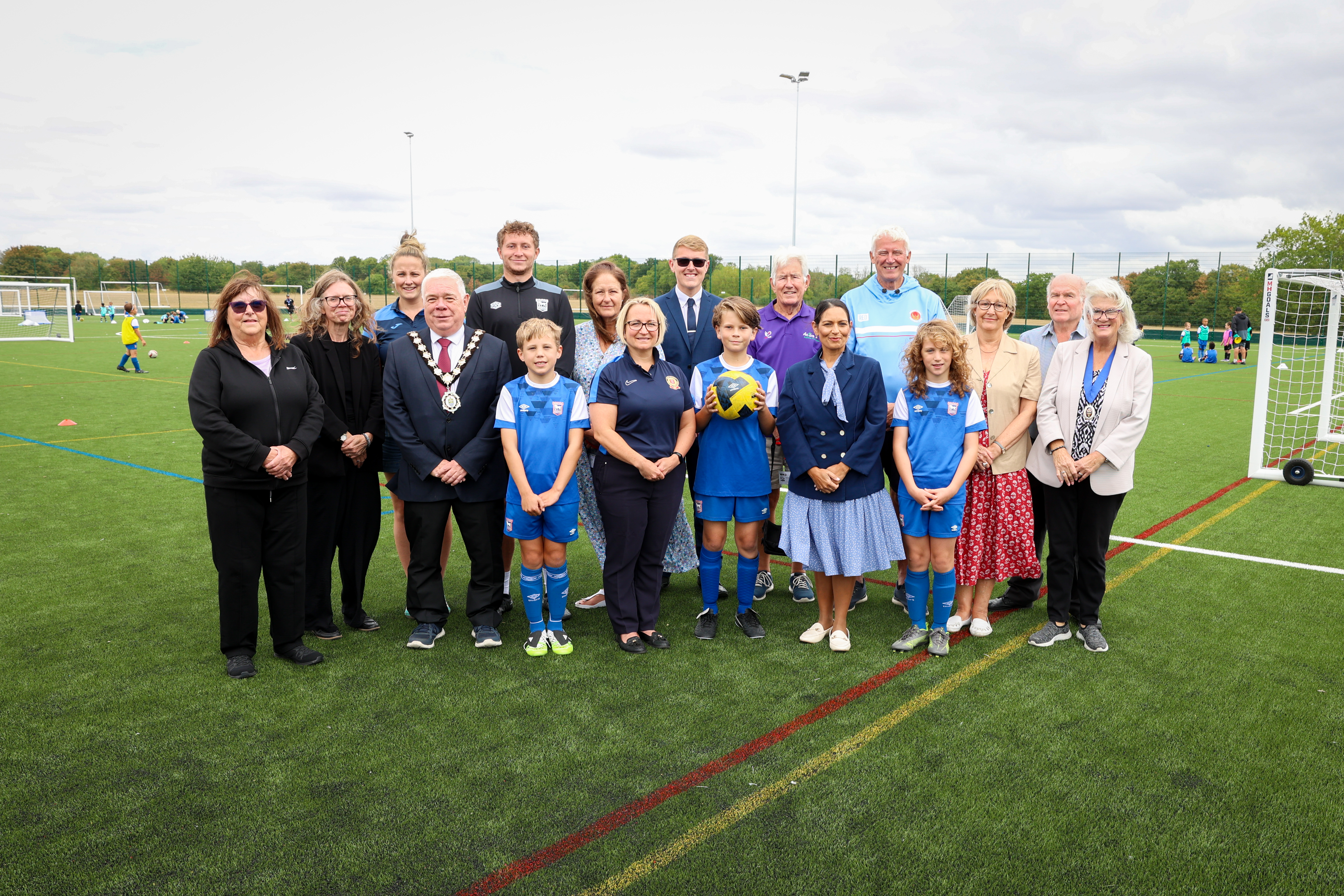 People smiling standing on artificial grass pitch in Witham with children playing football in the background